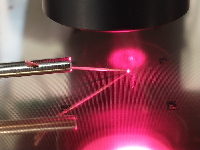 Probe needles contacting a VCSEL die under a microscope, with the emitted beam visible on the wafer